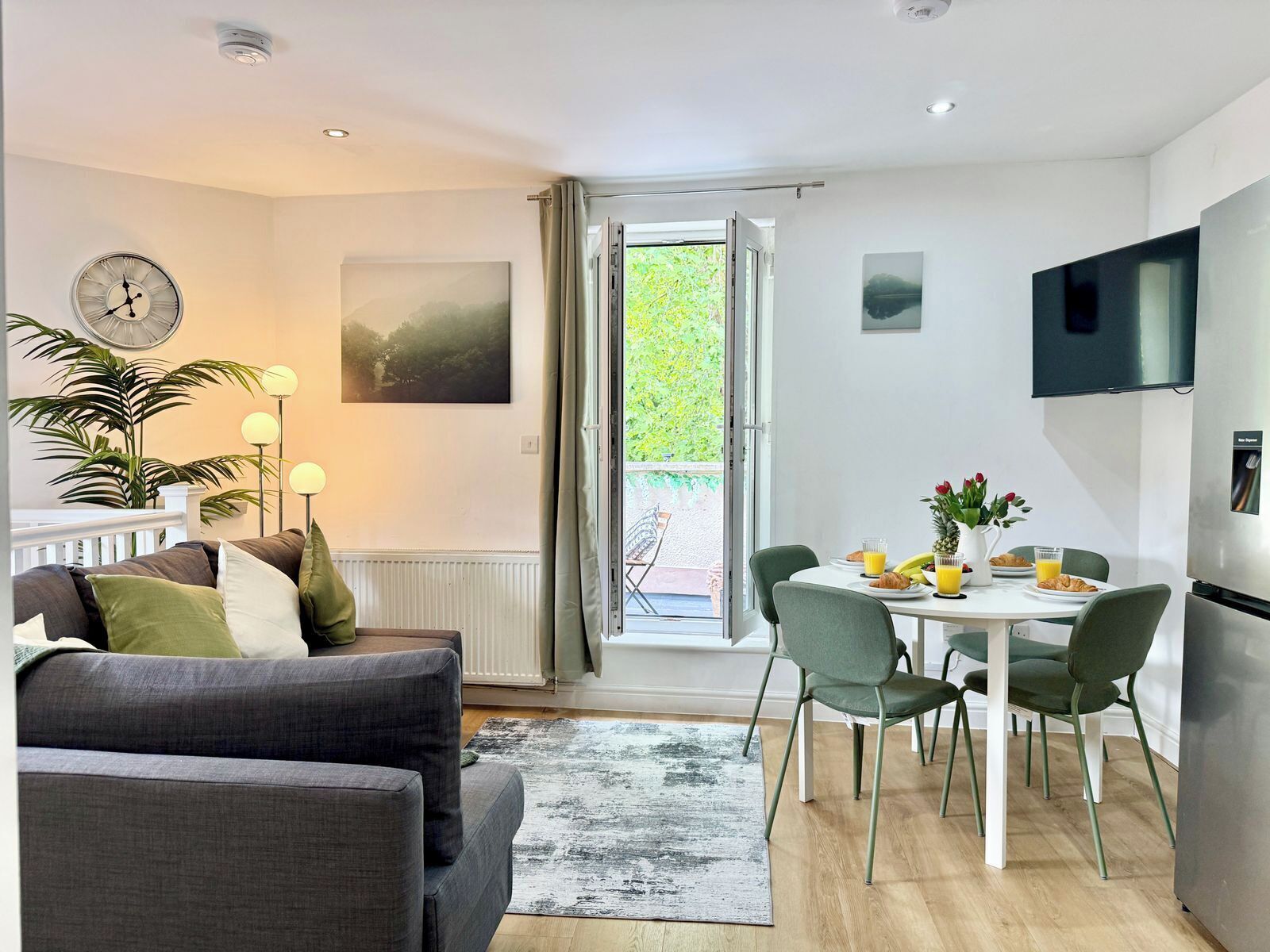 A modern living room with a sofa, a dining table with chairs, a clock, and a TV mounted on the wall.