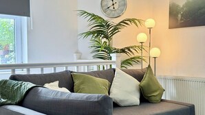Uma sala de estar com um sofá cinza, uma grande planta em vaso, um relógio de parede e um abajur.