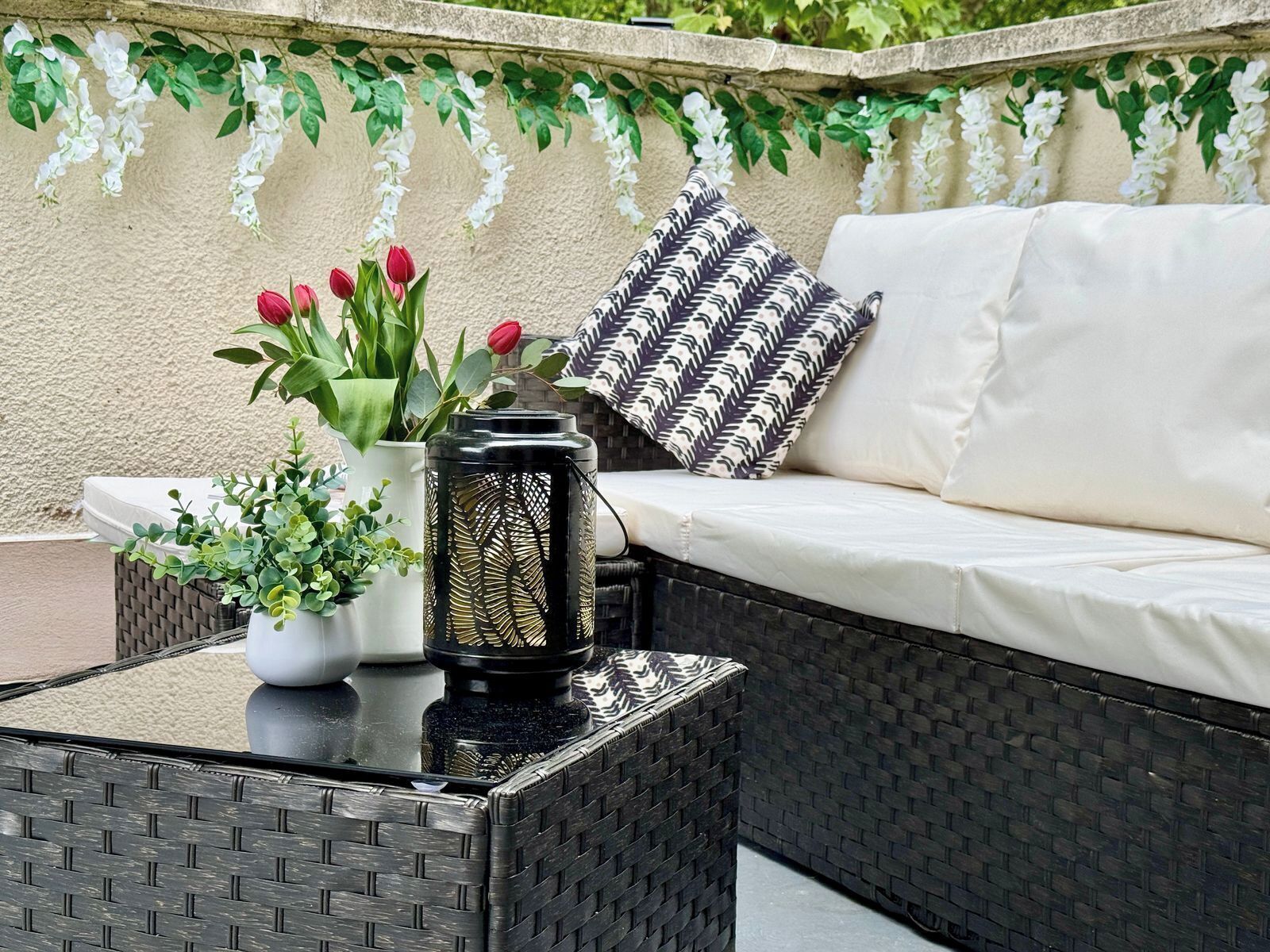 Un sofá de mimbre con cojines blancos, una almohada estampada en blanco y negro, una planta en maceta con tulipanes rojos y un farol decorativo.