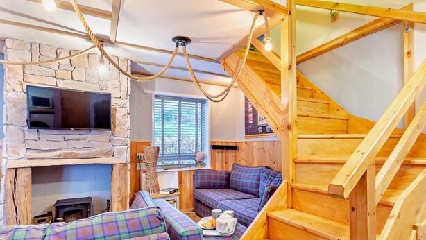 L’intérieur chaleureux d’un chalet avec une cheminée en pierre, un téléviseur, un escalier en bois et des sièges à motifs écossais.