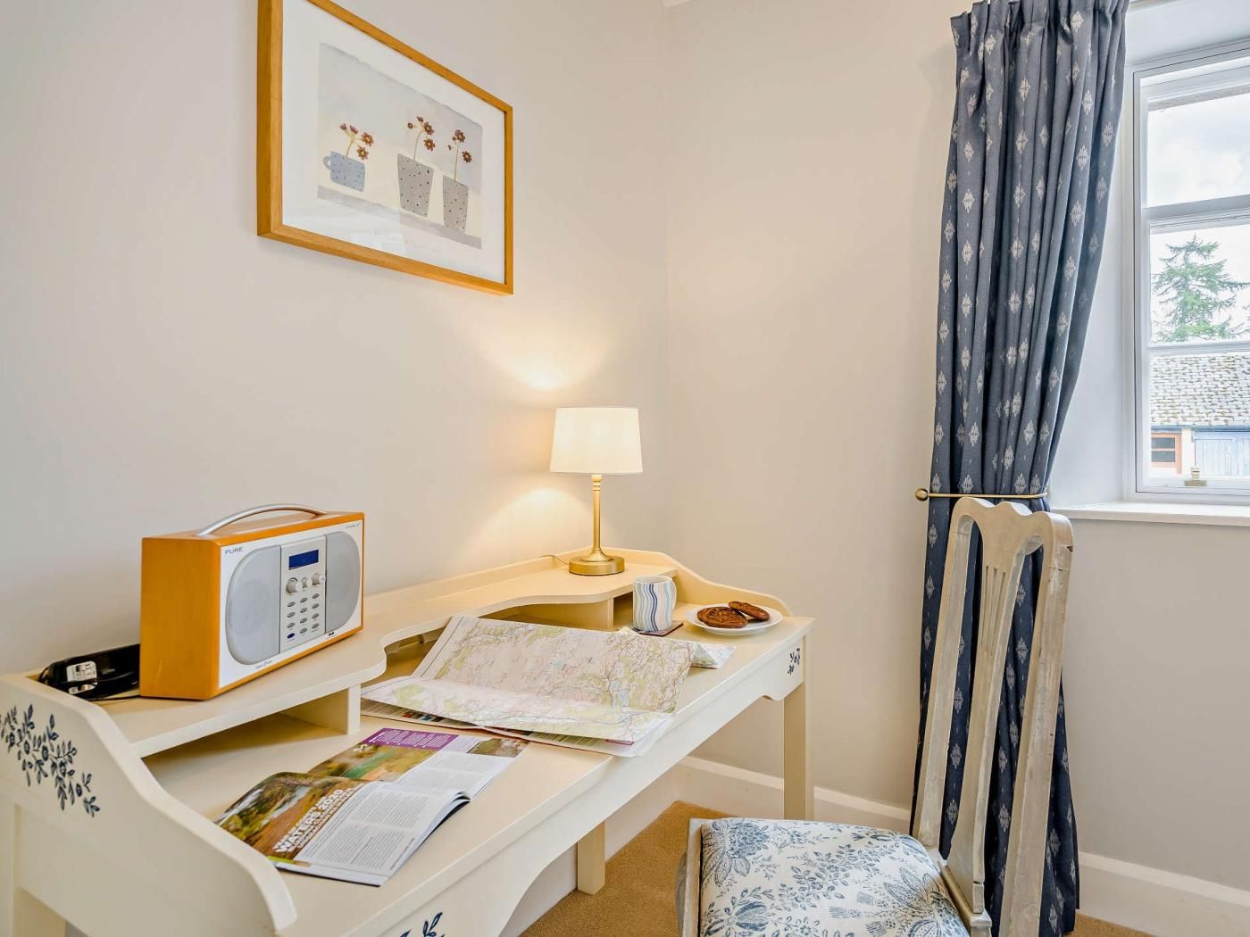 Et værelse med skrivebord, radio, lampe, stol, vindue med gardiner og et billede i ramme på væggen.