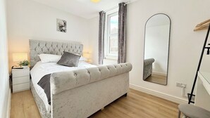 Un dormitorio con cama de cabecera capitonada, una mesita de noche con lámpara, un espejo grande y una ventana con cortinas.