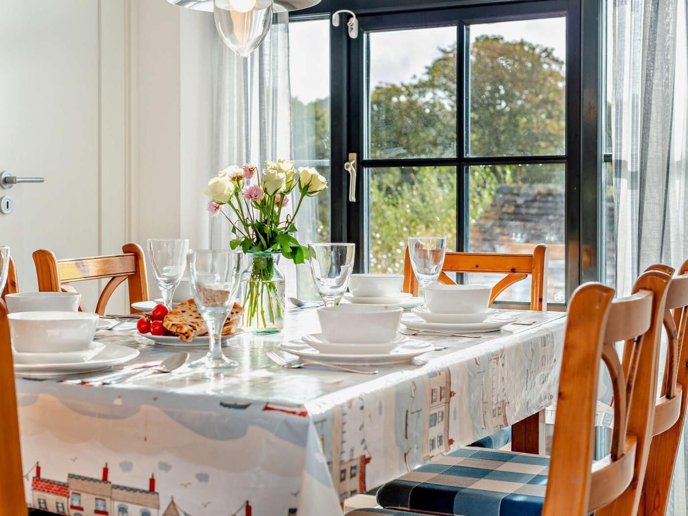 A dining table set with white plates, glasses, and a vase of flowers, with a view of trees through a large window.