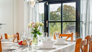 Ett matbord dukat med vita tallrikar, glas och en vas med blommor, med utsikt över träd genom ett stort fönster.