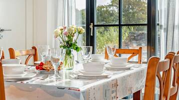 A dining table set with white plates, glasses, and a vase of flowers, with a view of trees through a large window.