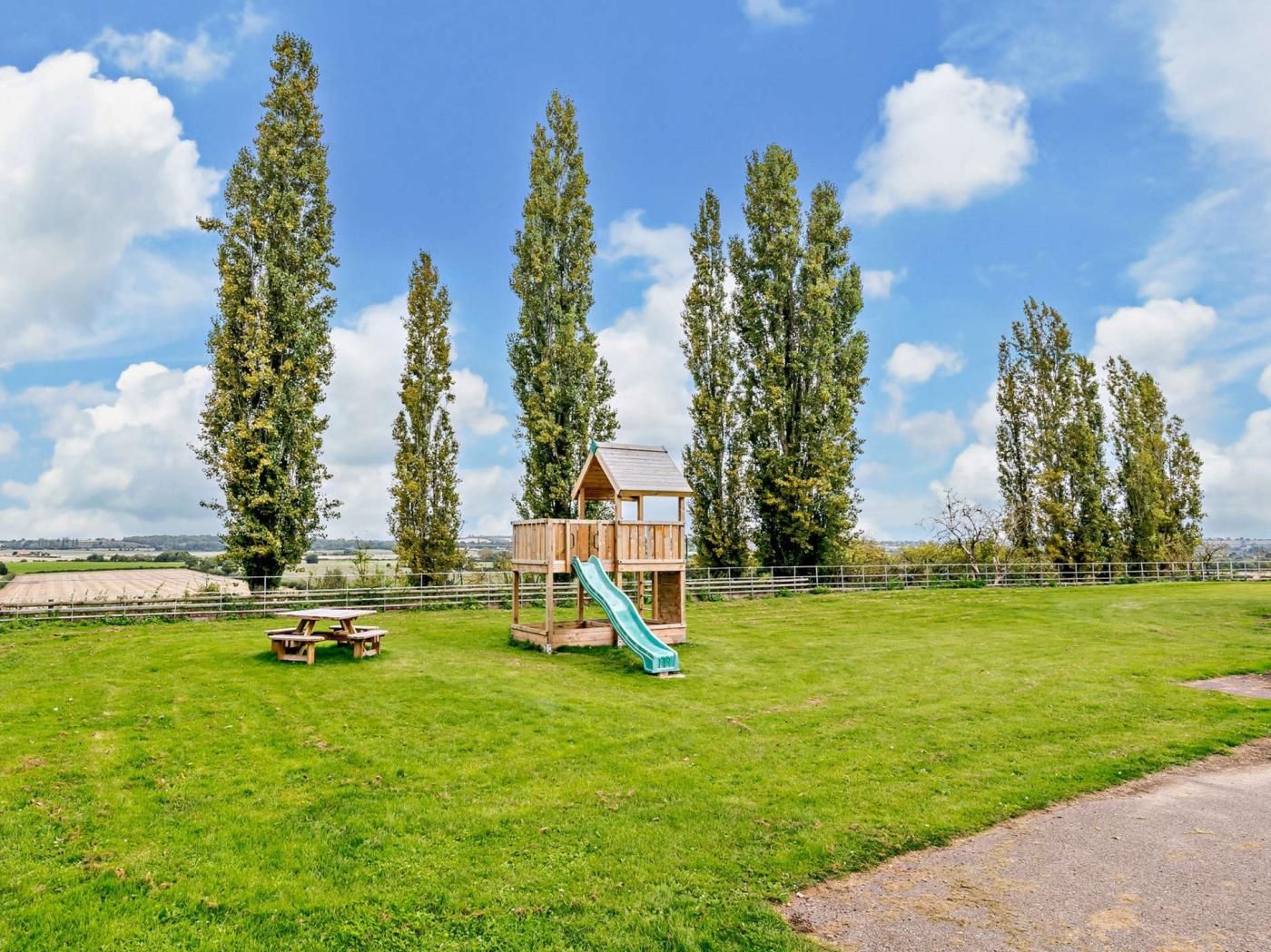 Een houten poppenhuis met een glijbaan in een grasveld, omringd door hoge bomen.