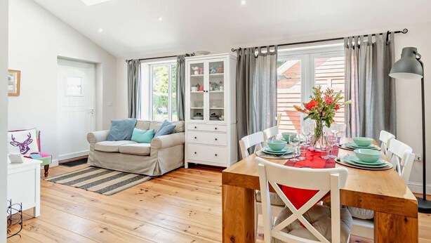 A modern, well-lit room with a wooden dining table, white chairs, a sofa with cushions, a cabinet, and a window with curtains. - 2 Bed in Driffield (oc-93140) (Langtoft)