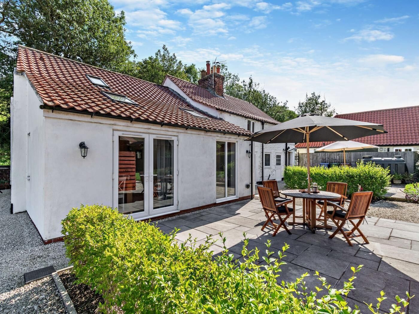 A small white house with a tiled roof, a patio with a table and chairs, and a large umbrella.