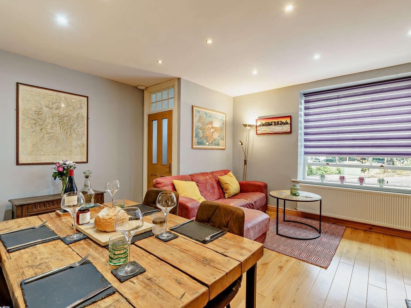 A cozy living room with a wooden dining table set for a meal, a red sofa, and a window with blinds.