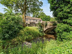 A stone bridge over a stream with lush greenery and a building in the background. - 2 Bed in Skipton (oc-92898) (Otterburn)