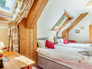 A bedroom with a sloped ceiling, wooden beams, a skylight, two beds with red and white bedding, a bedside table with a lamp, and a window with floral curtains. - 3 Bed in Weymouth (oc-92902) (Osmington)