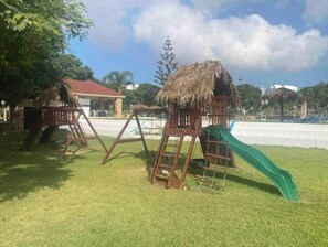 Un parque infantil con un tobogán y columpios, rodeado de césped y árboles.