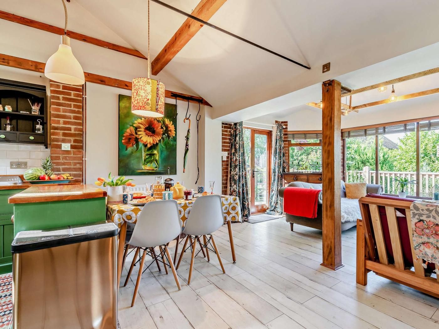 A spacious dining area with a wooden table and chairs, a kitchen with a brick wall, and a large window offering a view of greenery.
