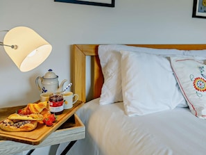 A neatly made bed with white linens and a decorative pillow, a bedside table with a tray of pastries and a teapot, and a wall-mounted lamp. - 3 Bed in Pershore (oc-92975) (Eckington)