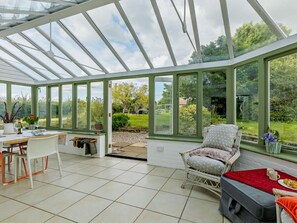 Ein Wintergarten mit Glasdach, einem Essbereich mit Tisch und Stühlen sowie einer Sitzecke mit Sessel und Kissen.