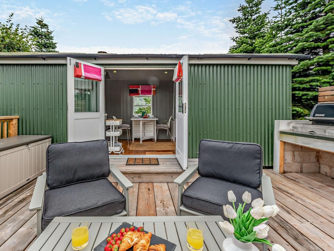 A cozy outdoor seating area with two chairs, a table with food and drinks, and a view of a green building with a door and windows.