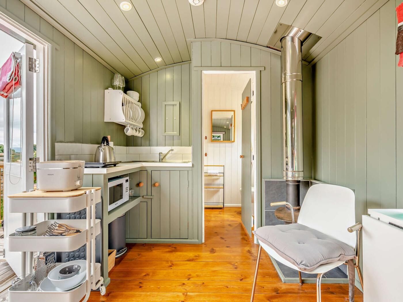 Una cocina compacta con microondas, hervidor y escurreplatos. Hay una silla blanca y una mesita con un hervidor.