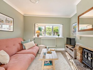 A living room with a sofa, a coffee table, a fireplace, a TV, and a window with a view of greenery. - 2 Bed in Bridport (oc-93076) (Burton Bradstock)