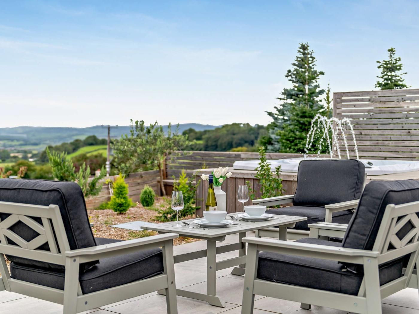 An outdoor patio with a table set for two, surrounded by chairs, a hot tub, and a wooden fence.
