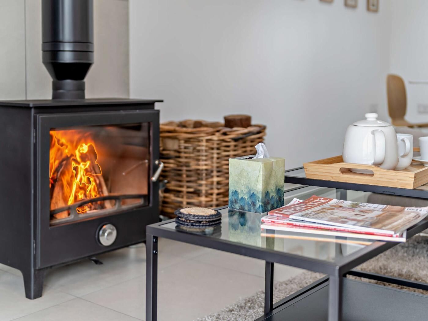 A modern living room with a wood-burning stove, a glass coffee table, a wicker basket, and a tea set.