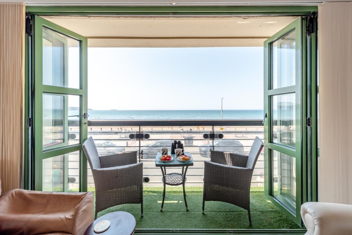 Un balcon avec des fauteuils en osier, une petite table sur laquelle est posé un plateau de nourriture, et une vue sur la plage.