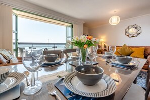 Ein Essbereich mit einem Tisch für zwei Personen, einer Blumenvase und Blick auf das Meer durch große Fenster.