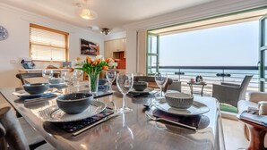 Een moderne eetruimte met uitzicht op de oceaan, een keuken op de achtergrond, en een eettafel gedekt met kommen, glazen en een vaas met bloemen.
