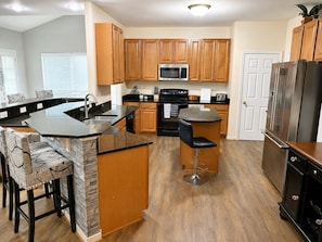 A kitchen with wooden cabinets, a central island with bar stools, a stainless steel refrigerator, and a microwave.