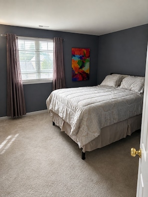 A bedroom with a bed, curtains, a window, and a colorful painting on the wall.