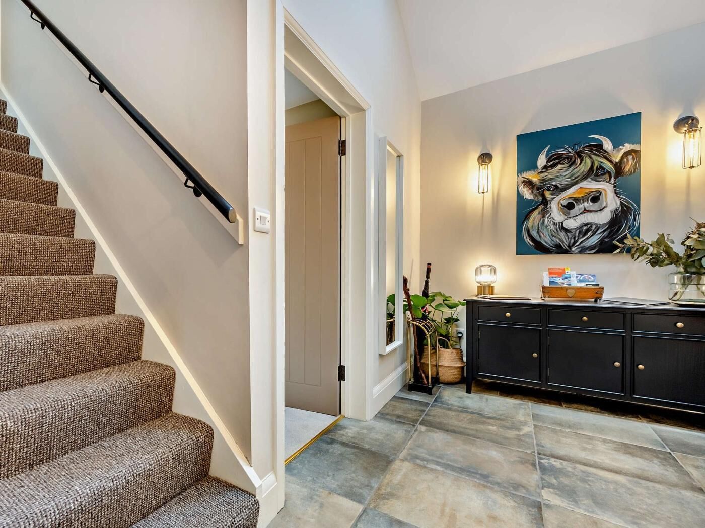 A staircase with a handrail, a hallway with a door, a wall-mounted painting, a cabinet with a lamp, and a potted plant.