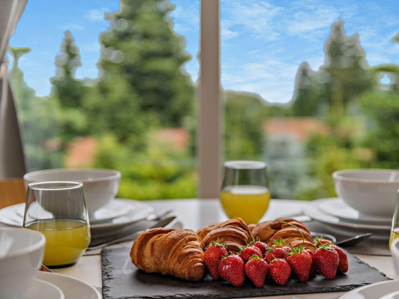 A breakfast setting with croissants and strawberries on a slate board, glasses of orange juice, and a view of greenery through a window.