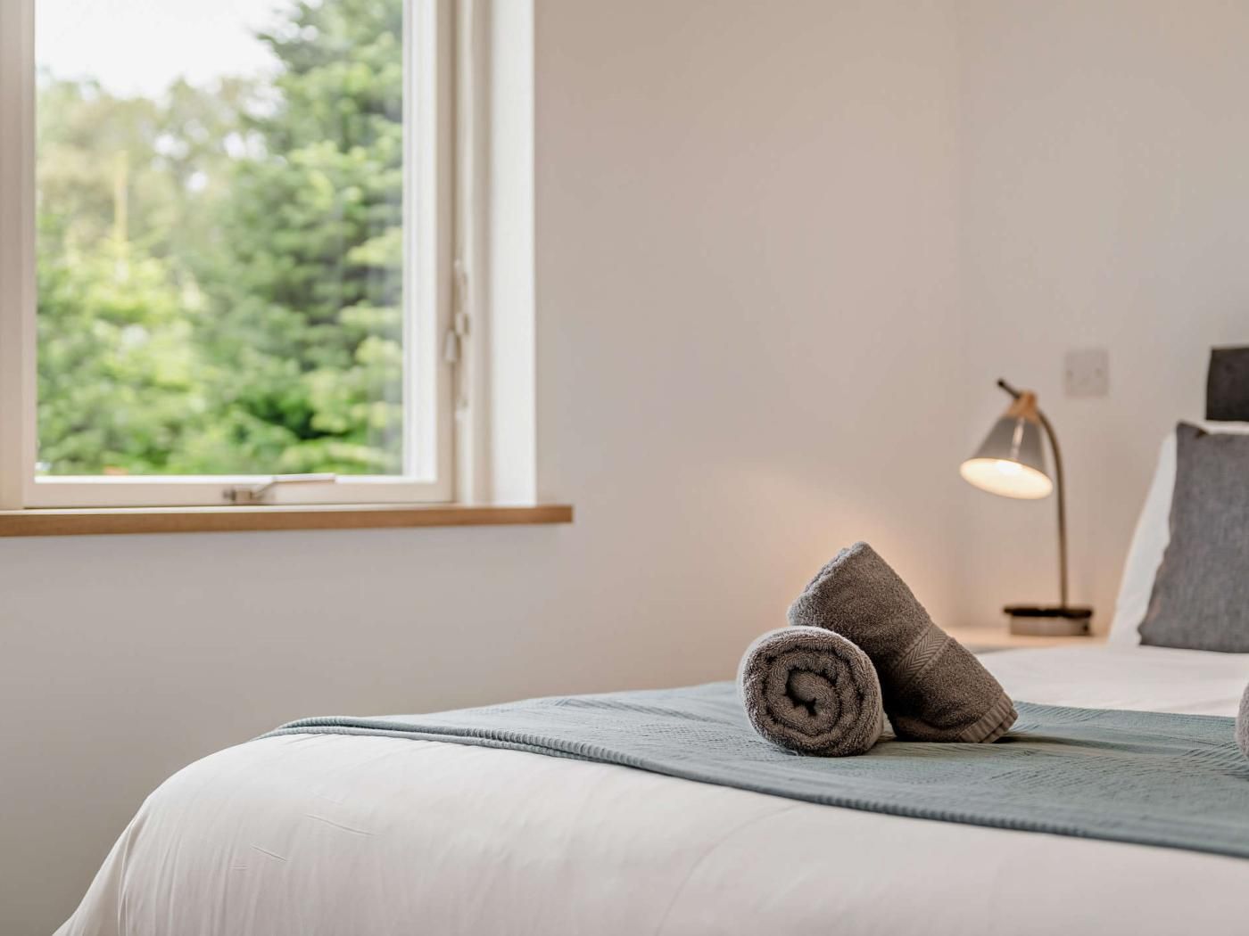A neatly made bed with a grey blanket and rolled towels, a bedside lamp, and a window with a view of trees.
