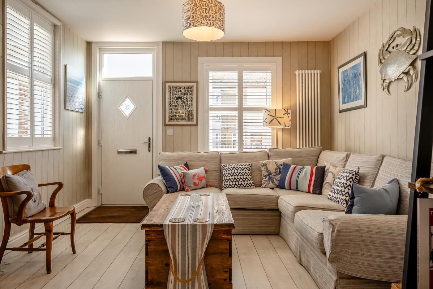 A cozy living room with a sofa, a wooden coffee table, and a chair. There is a door with a sign, a window with blinds, and a wall-mounted lamp.