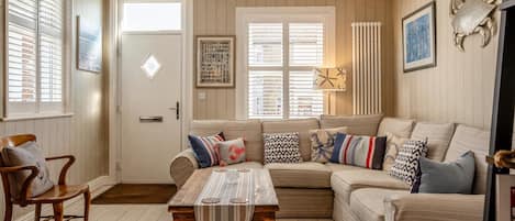A cozy living room with a sofa, a wooden coffee table, and a chair. There is a door with a sign, a window with blinds, and a wall-mounted lamp.