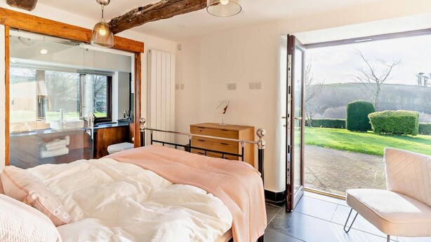 A bedroom with a large bed, a wooden headboard, a dresser, a mirror, and a view of a garden through a glass door. - 5 Bed in Broughton-in-furness (oc-93178) (Kirkby Ireleth)