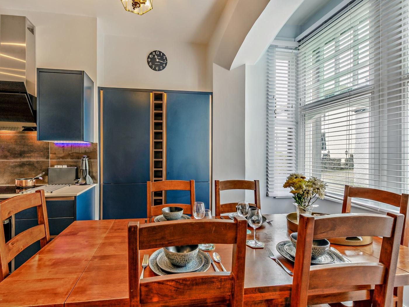 A dining area with a wooden table set for four, a clock on the wall, and a view of the outside through a window with blinds.