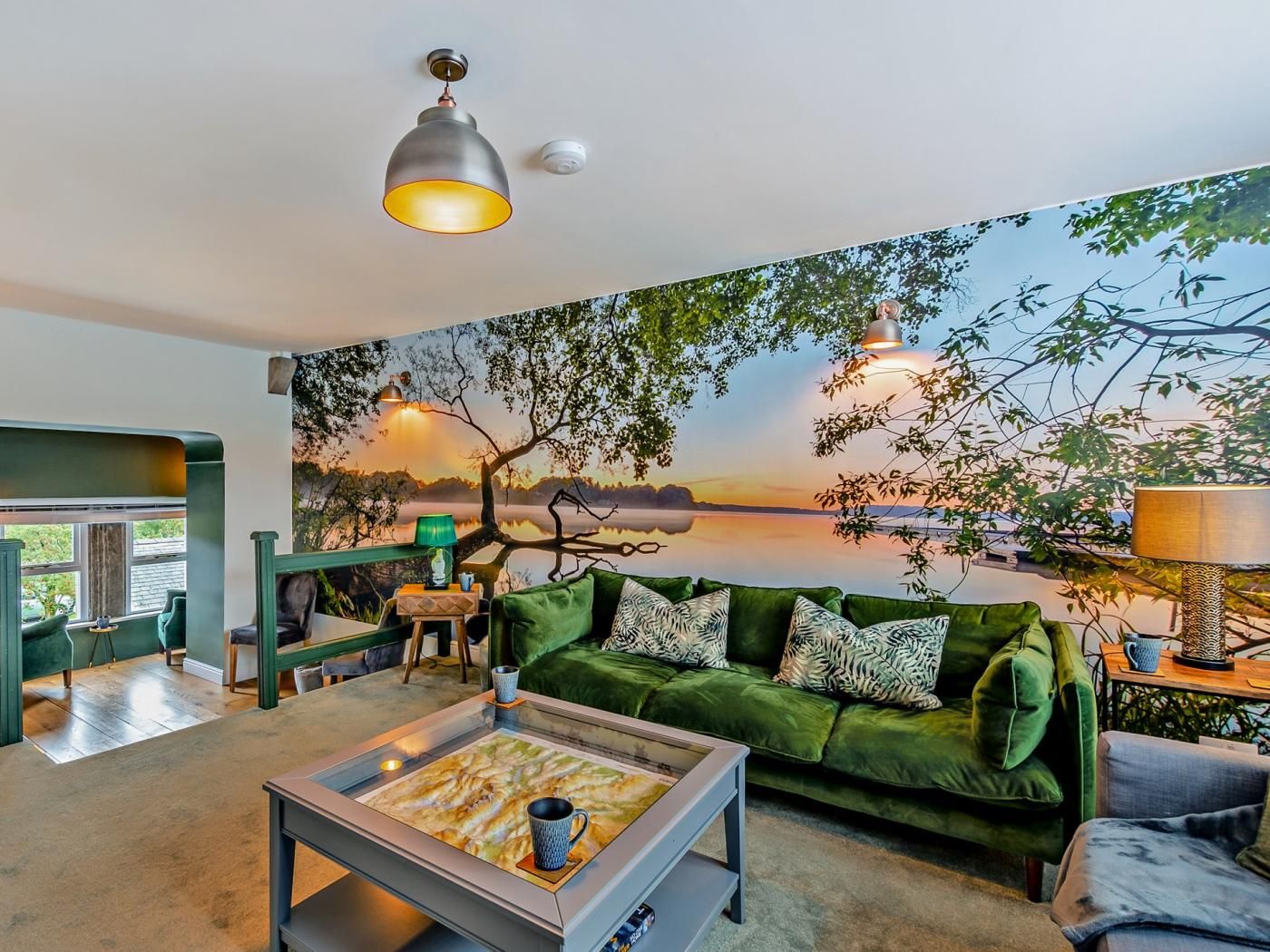 Un salon avec un canapé vert, une table basse et une fresque murale représentant un coucher de soleil sur un lac.