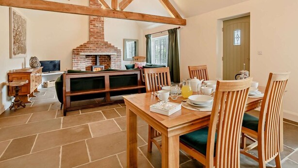 A rustic interior with exposed wooden beams, a brick fireplace, a dining table with chairs, and a television in the background. - 2 Bed in Harrogate (oc-92721) (Bishop Monkton)