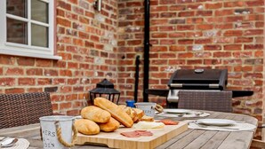 An outdoor dining area with a brick wall, a grill, and a wooden table set with bread, plates, and cutlery. - 2 Bed in Harrogate (oc-92721) (Bishop Monkton)