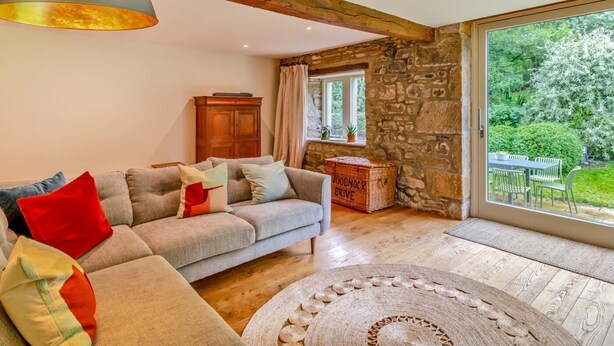 Un salon chaleureux avec un canapé, une poutre en bois au plafond, un mur en pierre et une vue sur le jardin.