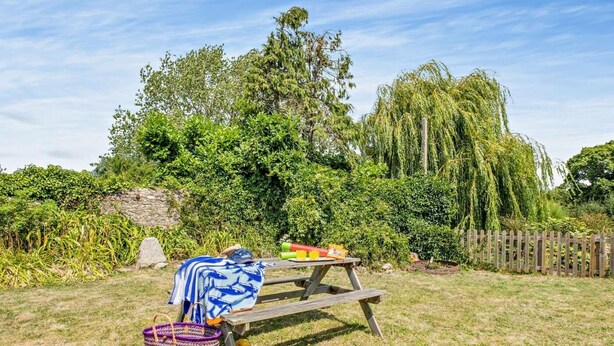 Ein Picknick mit Decke, Korb und Speisen auf einem Holztisch in einer grünen Umgebung mit Bäumen und einem Zaun.