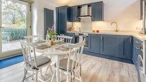 Uma cozinha moderna com armários azul escuro, uma mesa de jantar branca com cadeiras, e uma janela grande que oferece vista para o exterior.