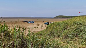 A beach with cars parked and people walking, grassy dunes in the foreground, and a clear sky. - 3 Bed in Burnham-on-sea (oc-92964) (Brean)