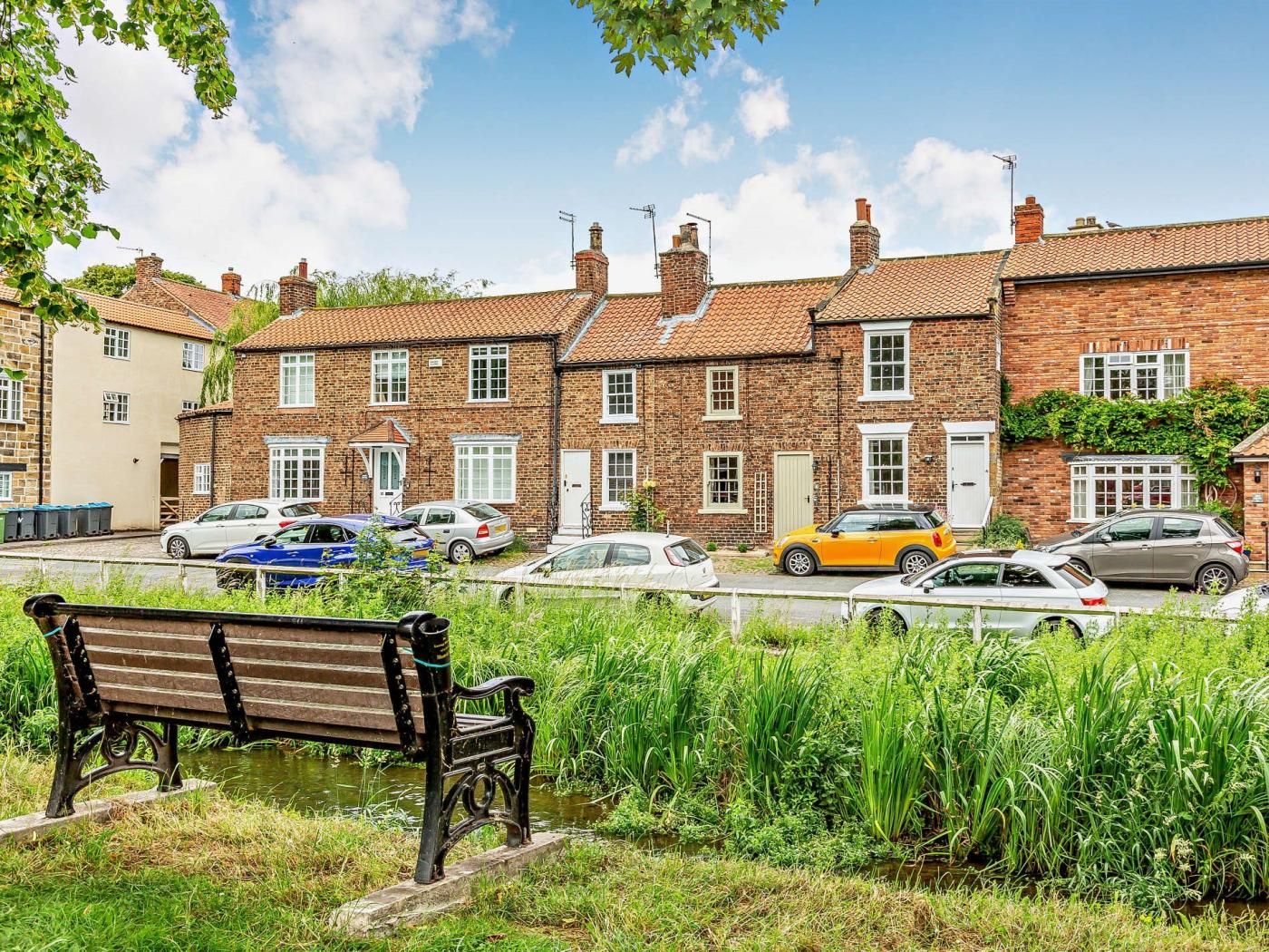 A row of brick houses with parked cars, a bench, and a waterway.