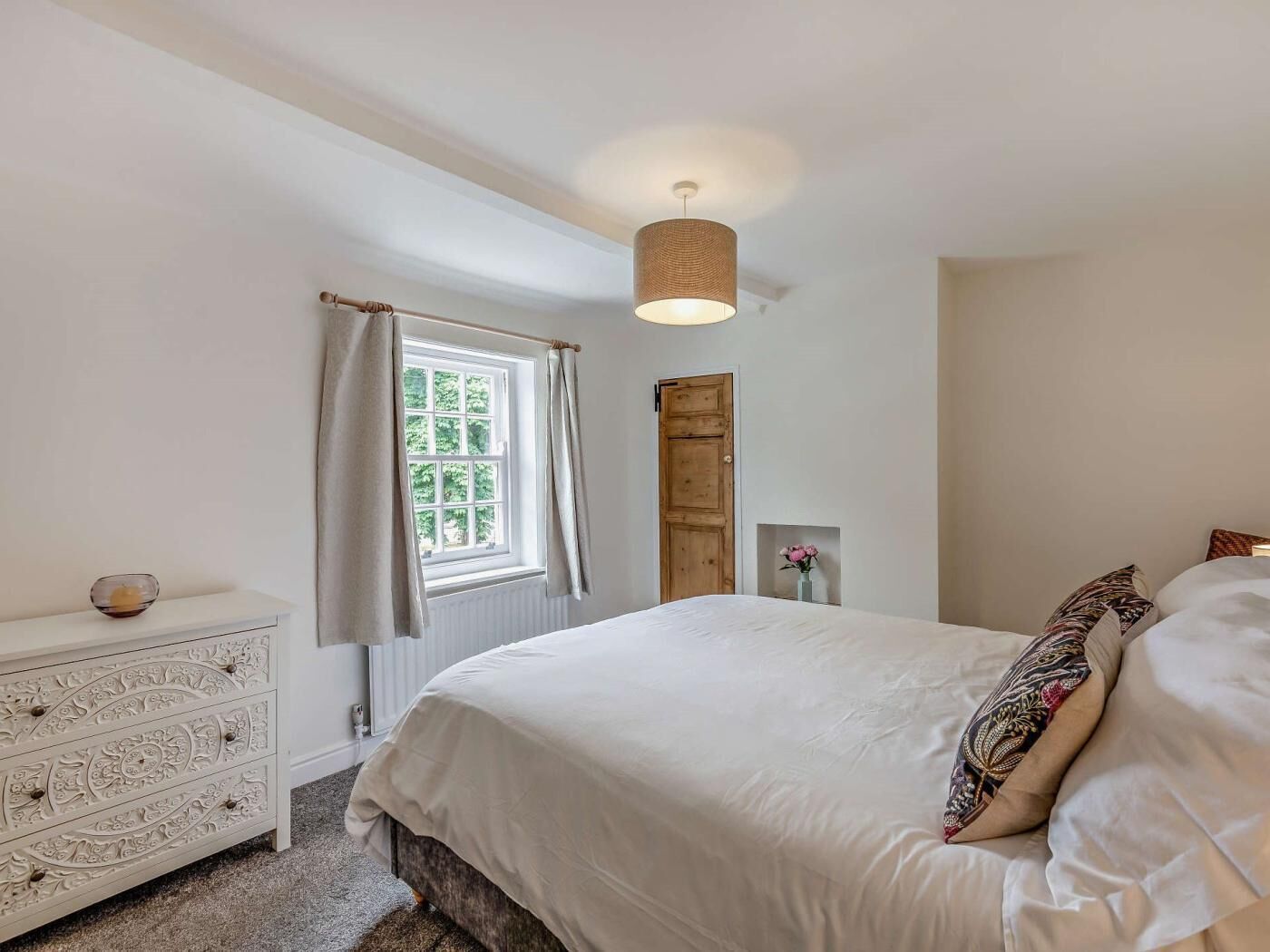 Un dormitorio con cama, cómoda, ventana con cortinas, puerta y lámpara colgante.