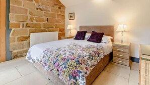 A bedroom with a stone wall, a wooden ceiling, a bed with floral bedding, two bedside lamps, and a small bedside table. - 1 Bed in Chop Gate (oc-93100) (Chop Gate)