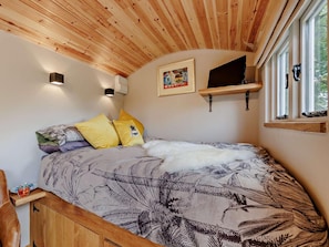 Ein Schlafzimmer mit Holzboden, einem Bett mit gemusterten Betttextilien, einem an der Wand befestigten Fernseher und einem Fenster mit Blick auf Grünflächen.