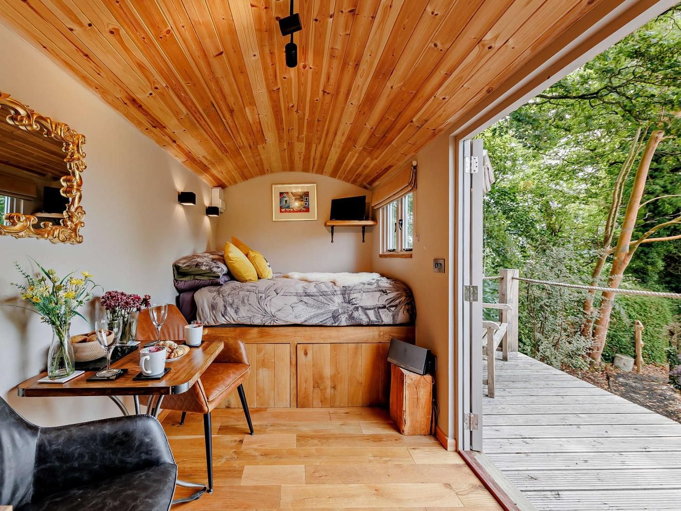 A cozy bedroom with a wooden ceiling, a bed with a floral bedspread, a desk with a chair, and a view of the outdoors.
