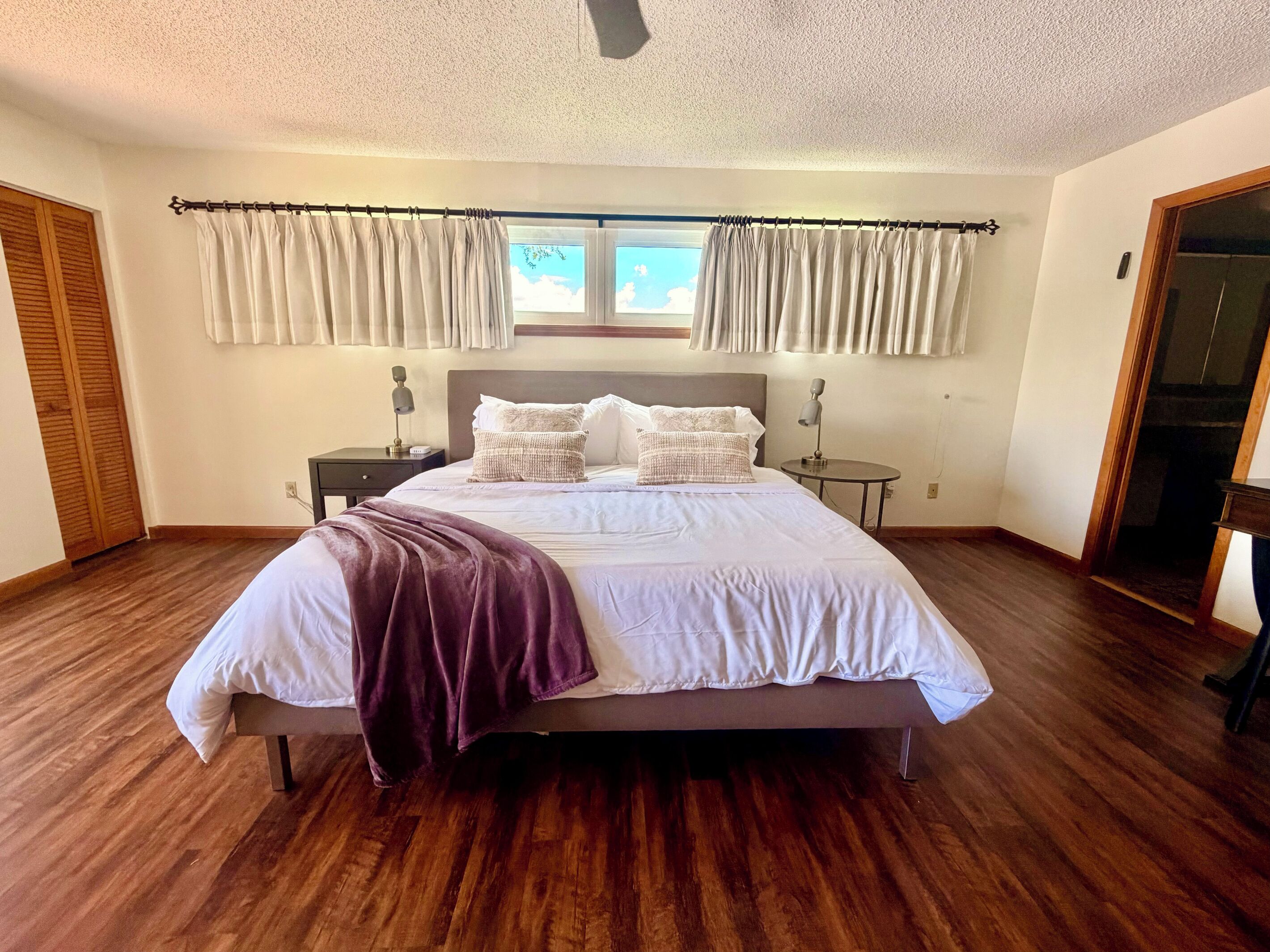 Une chambre à coucher avec un lit, deux tables de chevet, une fenêtre avec des rideaux, un plancher en bois et une porte.