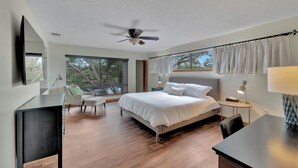 Ein modernes Schlafzimmer mit einem großen Bett, einem Schreibtisch, einem Stuhl, einem Deckenventilator und Blick auf Bäume durch das Fenster.